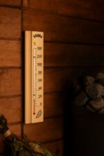 Koka pirts termometrs ar temperatūras skalu piestiprināts pie koka sienas blakus pirts krāsns akmeņiem