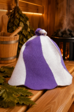 Balta un violeta filca pirts cepure "Ceriņi" novietota uz koka lāvas pirts interjerā. Fonā redzams pirts spainis, pirts slota un pirts krāsns.