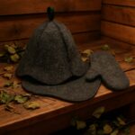 Pelēka filca pirts cepure un pirts dūrainis, novietoti uz koka lāvas pirts telpā