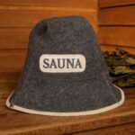 Tumši pelēka filca pirts cepure ar uzrakstu "SAUNA" priekšpusē, novietota uz koka lāvas pirts interjerā