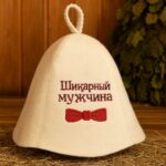 Gaiša filca pirts cepure ar sarkanu tauriņa aplikāciju un krievu uzrakstu “Шикарный мужчина” priekšpusē, novietota uz koka lāvas pirts interjerā