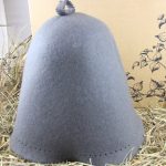 Pelēka filca pirts cepure ar dekoratīvu bumbuli augšpusē novietota uz salmiem pie gaišas sienas. Attēlā redzama cepures priekšpuse un gludā filca virsma.