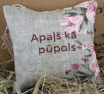 Aromātiskais spilventiņš ar uzrakstu "Apaļš kā pūpols" novietots uz salmiem pie gaiša fona. Attēlā izceļas dabīgā lina auduma faktūra, bordo krāsas uzraksts un rozā ziedu raksti spilventiņa malās.