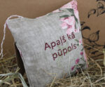 Aromātiskais spilventiņš ar uzrakstu "Apaļš kā pūpols" novietots uz salmiem un fotografēts no sānu leņķa. Attēlā redzams gaišs lina audums, rozā ziedu raksti un cilpiņa pakarināšanai.