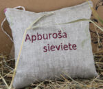 Aromātiskais spilventiņš ar uzrakstu "Apburoša sieviete" novietots uz salmiem pie gaiša fona. Attēlā redzama spilventiņa priekšpuse, dabīgā lina auduma faktūra un dekoratīvās zāles stiebrs pāri spilventiņam.