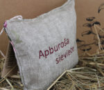 Aromātiskais spilventiņš ar uzrakstu "Apburoša sieviete" fotografēts no sāna un novietots uz salmiem. Attēlā izceļas dabīgā lina auduma faktūra, bordo krāsas uzraksts un dekoratīvā cilpiņa augšpusē.
