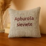 Aromātiskais auduma spilventiņš ar uzrakstu “Apburoša sieviete”, novietots uz koka virsmas