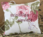 Aromātiskais spilventiņš ar uzrakstu "Brīnišķīga sieviete" novietots uz salmu fona. Uz spilventiņa redzami lieli rozā ziedu motīvi, gaišs lina audums un cilpiņa pakarināšanai.