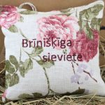 Aromātiskais spilventiņš ar uzrakstu "Brīnišķīga sieviete" novietots uz salmu fona. Uz spilventiņa redzami lieli rozā ziedu motīvi, gaišs lina audums un cilpiņa pakarināšanai.