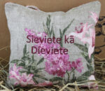 Aromātiskais spilventiņš ar uzrakstu "Sieviete kā Dieviete" novietots uz salmiem pie gaišas sienas. Uz lina auduma redzami rozā ziedu motīvi un aukliņa pakarināšanai.