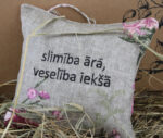 Dekoratīvs lina spilventiņš ar uzrakstu "slimība ārā, veselība iekšā" novietots uz salmiem pie gaišas sienas. Spilventiņam ir ziedu raksts un aukliņa pakarināšanai.