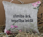 Dekoratīvs lina spilventiņš ar uzrakstu "slimība ārā, veselība iekšā" novietots uz salmiem pie gaišas sienas. Uz spilventiņa redzami ziedu motīvi un aukliņa pakarināšanai.