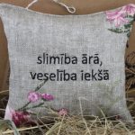 Dekoratīvs lina spilventiņš ar uzrakstu "slimība ārā, veselība iekšā" novietots uz salmiem pie gaišas sienas. Uz spilventiņa redzami ziedu motīvi un aukliņa pakarināšanai.