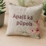 Aromātiskais auduma spilventiņš ar uzrakstu “Apaļš kā pūpols”, novietots uz gaišas koka virsmas