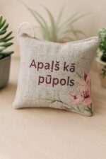 Aromātiskais auduma spilventiņš ar uzrakstu “Apaļš kā pūpols”, novietots uz gaišas koka virsmas