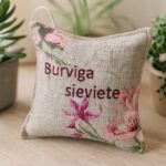 Aromātiskais auduma spilventiņš ar uzrakstu “Burvīga sieviete”, novietots uz gaišas koka virsmas