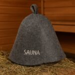 Pelēka filca pirts cepure ar uzrakstu SAUNA, novietota uz salmiem koka pirts interjerā