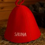 Sarkana filca pirts cepure ar baltu uzrakstu “SAUNA”, novietota uz koka lāvas tradicionālā pirts interjerā