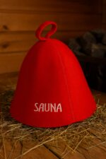 Sarkana filca pirts cepure ar baltu uzrakstu “SAUNA”, novietota uz koka lāvas tradicionālā pirts interjerā