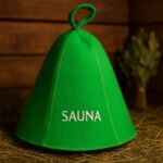 Zaļa filca pirts cepure ar baltu uzrakstu “SAUNA”, novietota uz koka lāvas tradicionālā pirts interjerā