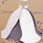 Balta un pelēka filca pirts cepure uz salmu fona. Attēlā redzama pirts cepures aizmugure ar kontrastējošu filca apdari un cilpu pakarināšanai.