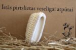 Ovāla pirts švammīte ar baltu mīkstu ielaidumu novietota vertikāli uz siena fona. Attēlā redzama švammītes sānu daļa, kas izceļ dabīgās lufas virsmu un mīksto tekstila pusi.