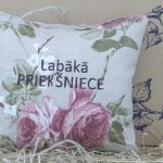 Aromātiskais spilventiņš ar uzrakstu "Labākā priekšniece" un rozā ziedu rakstu novietots uz siena fona. Attēlā redzams dekoratīvs kvadrātveida spilventiņš gaišos toņos ar piekariņu augšpusē.