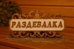 Koka dekoratīva pirts izkārtne ar uzrakstu “Раздевалка”, novietota uz salmiem uz lāvas