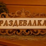 Koka dekoratīva pirts izkārtne ar uzrakstu “Раздевалка”, novietota uz salmiem uz lāvas