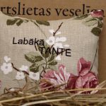 Aromātiskais spilventiņš ar uzrakstu "Labākā tante" un ziedu rakstu novietots uz siena fona. Attēlā redzams dekoratīvais spilventiņš ar rozā ziediem un izšūtu uzrakstu.