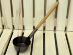 Melns alumīnija pirts kauss ar garu koka rokturi novietots diagonāli uz gaišām koka lāvām. Attēlā izcelts kausa augšskats un roktura novietojums.