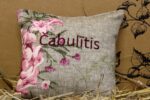 Aromātiskais spilventiņš ar uzrakstu "Cābulītis" novietots uz siena fona. Attēlā redzams gaišs auduma spilventiņš ar rozā ziedu rakstiem un izšūtu uzrakstu.