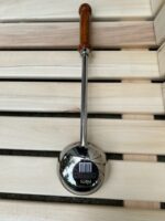 Nerūsējošā tērauda pirts kauss ar koka rokturi novietots uz gaišām koka lāvām. Attēlā redzams tradicionāls pirts kauss ar spīdīgu metāla virsmu un garu koka rokturi.