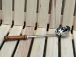 Nerūsējošā tērauda pirts kauss ar koka rokturi novietots uz gaišām koka lāvām. Attēlā redzams tradicionāls pirts kauss ar spīdīgu metāla virsmu un ergonomisku koka rokturi.