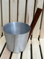 Alumīnija pirts ķipis ar koka rokturi novietots uz gaišām koka lāvām. Attēlā redzams viegls alumīnija ķipis ar dabīga koka rokturi tradicionālai lietošanai pirtī.
