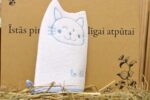 Balta kaķa formas bērnu vannas švammīte novietota vertikāli uz salmu fona. Attēlā redzams sānskats ar zilu kaķa sejas izšuvumu un gaiši zilu apmali švammītes apakšdaļā.