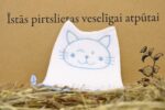 Balta bērnu pirts cepure ar zilu kaķa sejas izšuvumu novietota salmos uz dekoratīva fona. Attēlā cepure fotografēta no priekšpuses, izceļot kaķa sejas zīmējumu un augsto smailo formu.