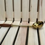 Nerūsējošā tērauda pirts kauss ar koka rokturi novietots uz gaišām koka lāvām. Attēlā redzams tradicionāls pirts piederums ūdens uzliešanai uz pirts akmeņiem.