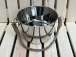 Nerūsējošā tērauda pirts ķipis ar dekoratīvu virves rokturi novietots uz gaišām koka lāvām. Attēlā redzams spīdīgs metāla ķipis tradicionālam pirts rituālam.