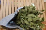 Pelēka filca pirts soma piepildīta ar lauru lapu pirts slotu novietota uz salmu fona pie koka lāvām. Attēlā izcelta ietilpīga pirts soma un aromātiskā lauru lapu slota.