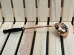 Vara pirts kauss ar melnu rokturi novietots uz gaišām koka lāvām. Attēlā redzams tradicionāls pirts kauss ar vara krāsas metāla daļu un ergonomisku rokturi.