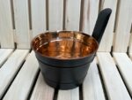 Vara pirts ķipis ar melniem rokturiem novietots uz gaišām koka lāvām. Attēlā redzams tradicionāls pirts ķipis ar spīdīgu vara iekšpusi un melnu ārējo apdari.