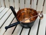Vara pirts ķipis ar melniem rokturiem un vara pirts kausu novietots uz gaišām koka lāvām. Attēlā redzams tradicionāls pirts komplekts ar spīdīgu vara apdari.