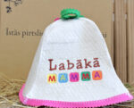 Balta filca pirts cepure ar uzrakstu "Labākā MAMMA" novietota uz salmu fona. Attēlā redzama pirts cepure ar krāsainu izšuvumu un rozā apdari ap malu.