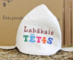 Balta filca pirts cepure ar uzrakstu "Labākais TĒTIS" novietota uz salmu fona. Attēlā redzama klasiska pirts cepure ar krāsainu izšuvumu priekšpusē.
