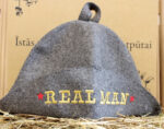 Pelēka filca pirts cepure ar uzrakstu "REAL MAN" novietota uz salmu fona. Attēlā redzama klasiska pirts cepure ar zelta krāsas izšuvumu un sarkanām zvaigznēm abās pusēs.