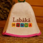 Gaiša filca pirts cepure ar uzrakstu “Labākā MAMMA” un zaļu cilpu augšpusē, ar rozā apmali apakšdaļā, novietota uz salmiem koka pirts interjerā