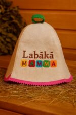 Gaiša filca pirts cepure ar uzrakstu “Labākā MAMMA” un zaļu cilpu augšpusē, ar rozā apmali apakšdaļā, novietota uz salmiem koka pirts interjerā