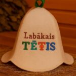 Gaiša filca pirts cepure ar uzrakstu “Labākais TĒTIS” un zaļu cilpu augšpusē, novietota uz salmiem koka pirts interjerā