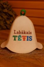 Gaiša filca pirts cepure ar uzrakstu “Labākais TĒTIS” un zaļu cilpu augšpusē, novietota uz salmiem koka pirts interjerā